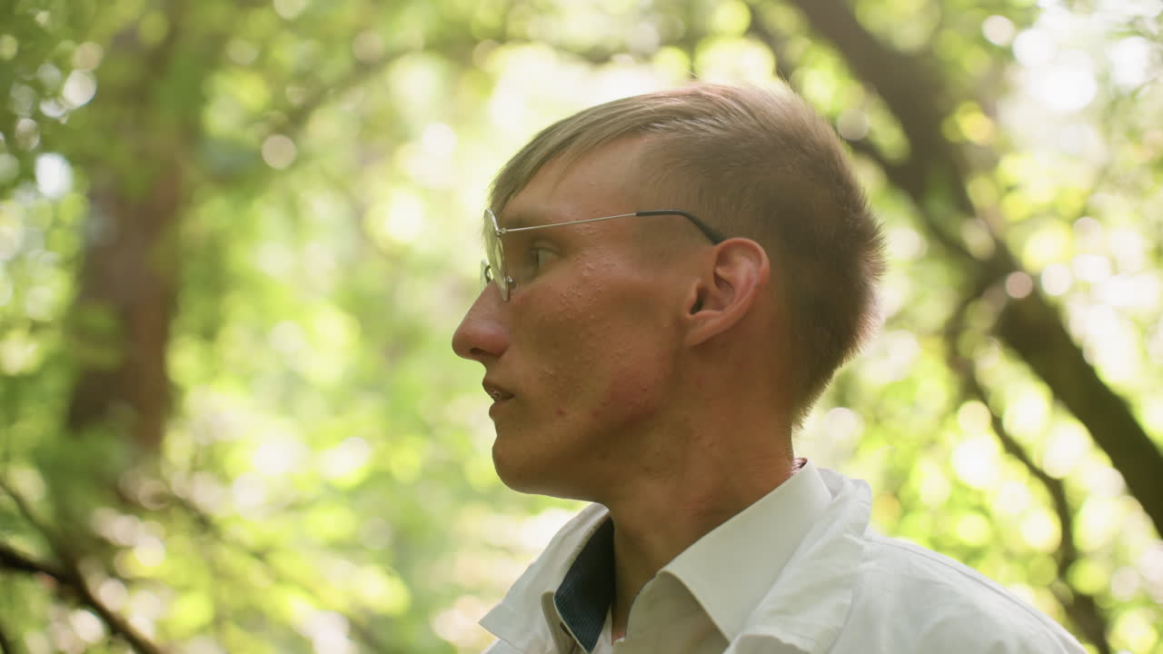 Young man wearing glasses adjusting white lab coat while standing in blurred forest environment with soft daylight creating natural background atmosphere highlighting outdoor research preparation