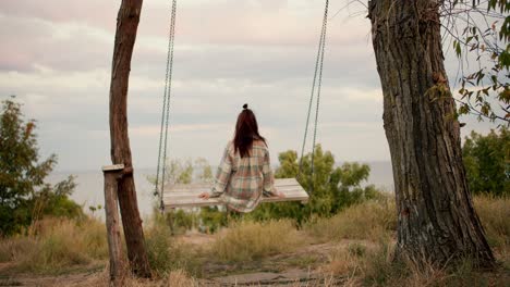 Shooting-from-behind:-A-brunette-girl-in-a-checkered-shirt-swings-on-a-wooden-swing-outside-the-city.-Rest-in-the-country-by-the-sea