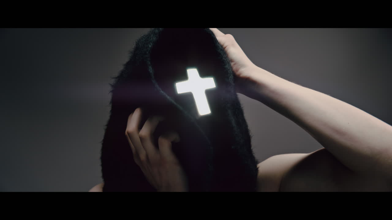 Masked man with cross of light glowing on his face grabs his head with his hand