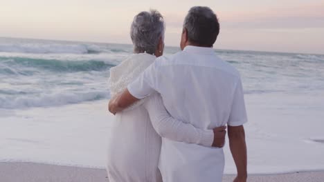 Rückansicht-Eines-Glücklichen-Hispanischen-Frisch-Verheirateten-älteren-Paares,-Das-Sich-Bei-Sonnenuntergang-Am-Strand-Umarmt