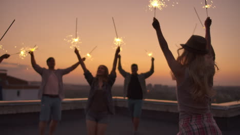 Young-people-raise-their-hands-up-and-jump-on-the-roof-with-big-bengal-light-in-their-hands.-It's-a-crazy-party-at-night.