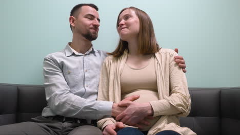 front view of pregnant woman and her husband sitting on sofa talking and waiting for gynecologist in medical consultation 2