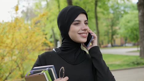 young arabic beautiful girl in hijab talking by mobile phone holding textbooks, glasses having nice appearance good mood. pretty muslim female student holding books, standing in the park near academy.