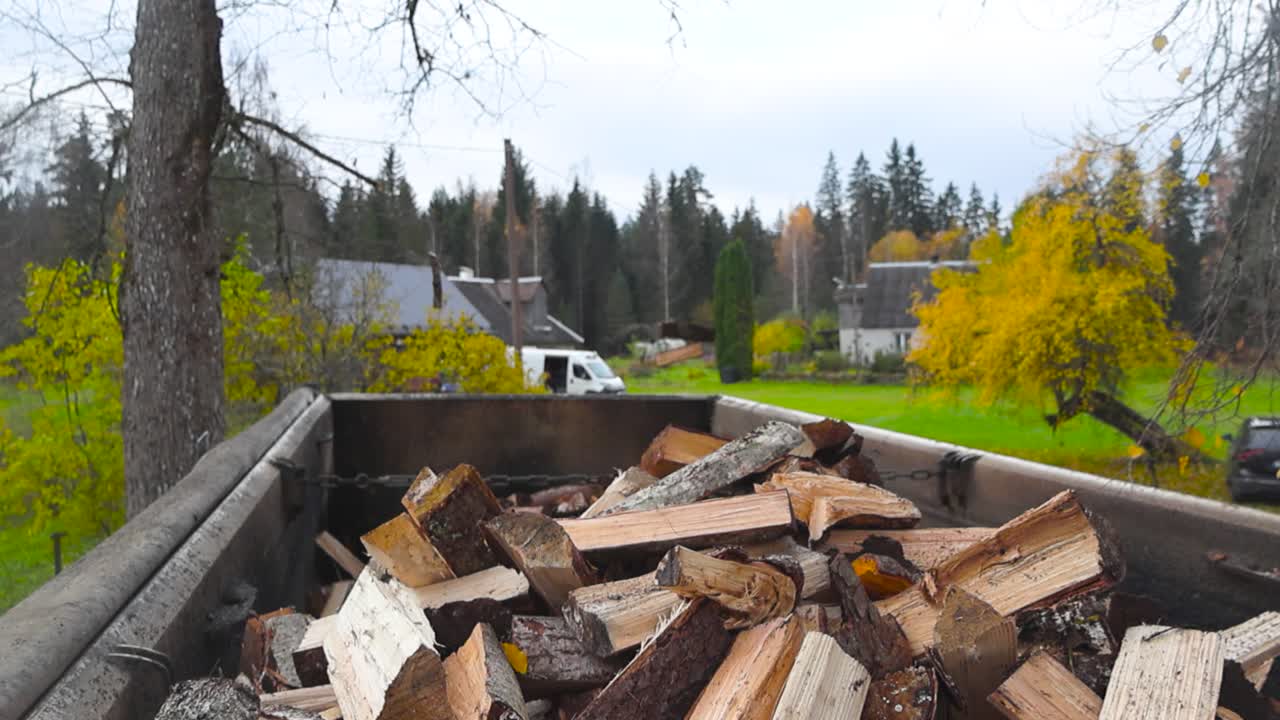 Cut and broken up firewood being thrown into a dump truck in slow motion during autumn cloudy day while trees have yellow leaves in the background garden. Timber is flying in the air smoothly.