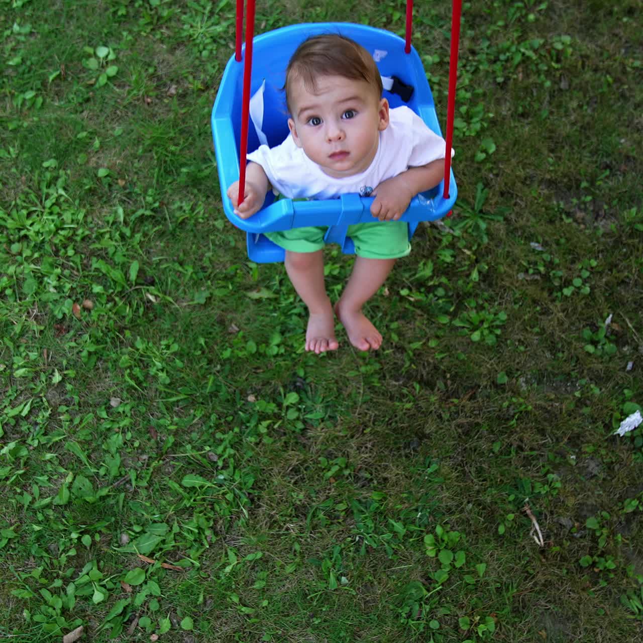 Curious small kid swaying in the swing. Baby looks up at camera. View from above. Green grass at backdrop