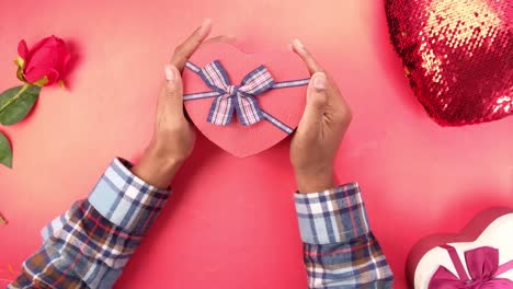 manos sosteniendo una caja de regalos en forma de corazón sobre un fondo rosa