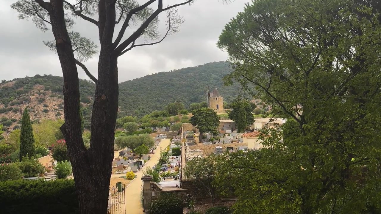 Un tranquillo cimitero collinare e una storica torre in pietra si trovano sotto montagne boscose, incorniciati da alberi alti sotto un cielo mediterraneo coperto