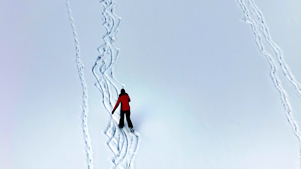 A lone skier moves across untouched snow in a red jacket, carving winding trails through a white landscape during a quiet winter day, viewed from above.