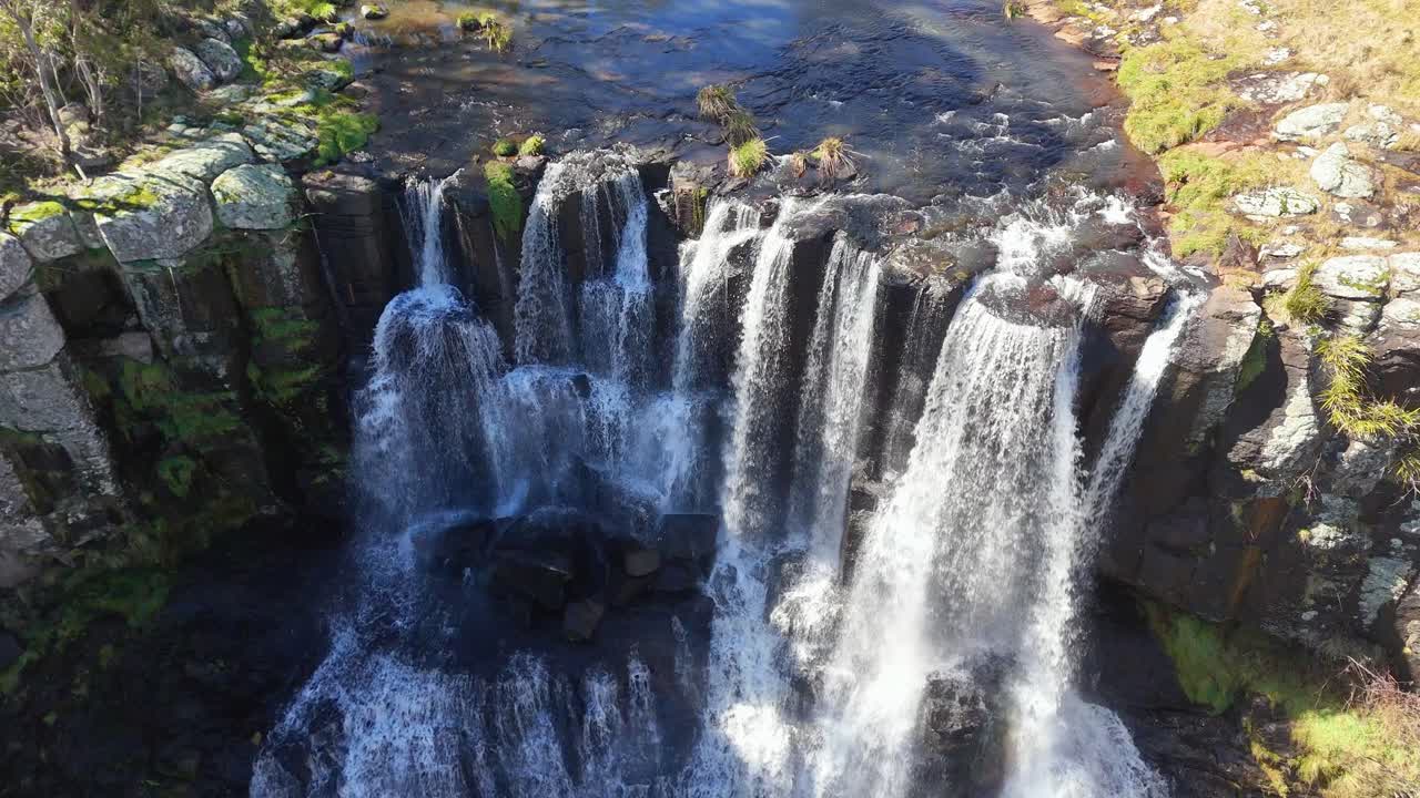 Drone footage captures Ebor Falls in New South Wales, Australia, with multiple streams of water plunging over mossy rocks in bright daylight