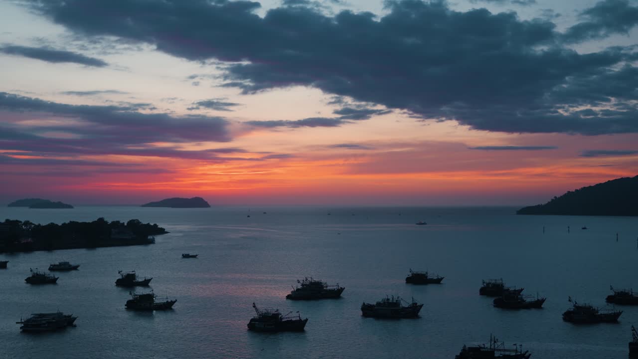 Scenic aerial panorama of Kota Kinabalu harbour at sunset shows a fleet of silhouetted fishing boats on calm sea with a dramatic orange sky and distant islands