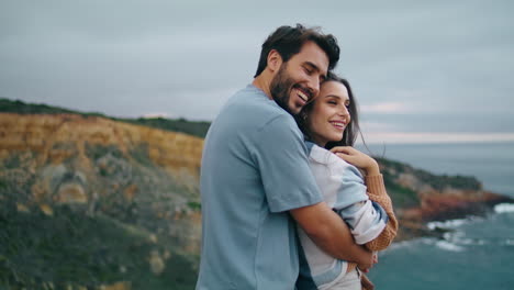 Smiling-pair-looking-camera-standing-hill-top-in-front-gray-calm-ocean-close-up.