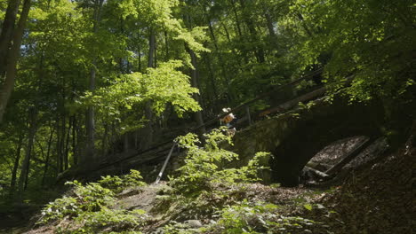 Young-girl-with-smartphone-and-gimbal-walking-on-bridge-in-green-sunny-landscape-of-Bohemian-Switzerland-National-Park,-Czech-Republic,-wide-view