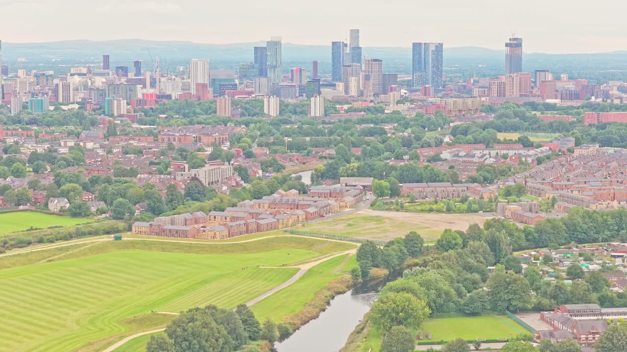 The River Irwell winds past green fields and new housing in Salford, with the modern Manchester skyline rising in the distance, captured in an ascending aerial drone shot