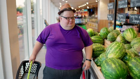 An-overweight-man-wearing-a-purple-T-shirt-and-a-brown-hat-walks-along-a-row-of-watermelons-in-a-large-supermarket-and-looks-at-them