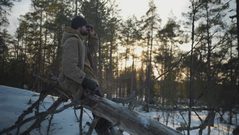 hombre sentado en un árbol caído y mirando a través de binoculares en la temporada de invierno