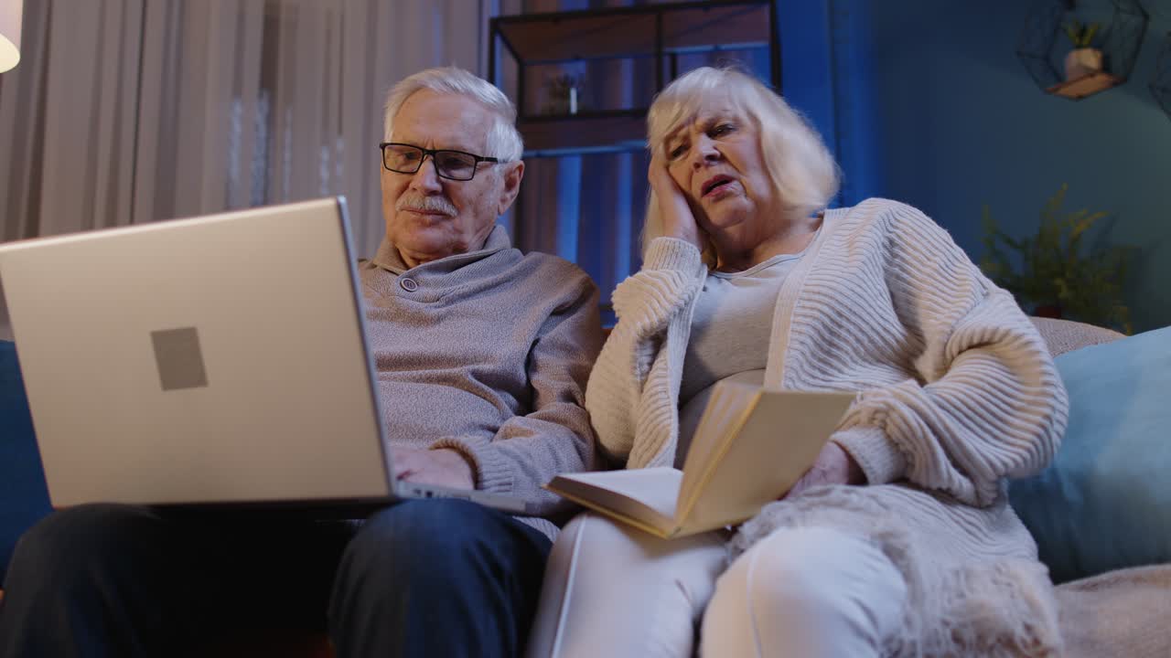Senior grandparents couple reading book using laptop pc on couch in night living room at home