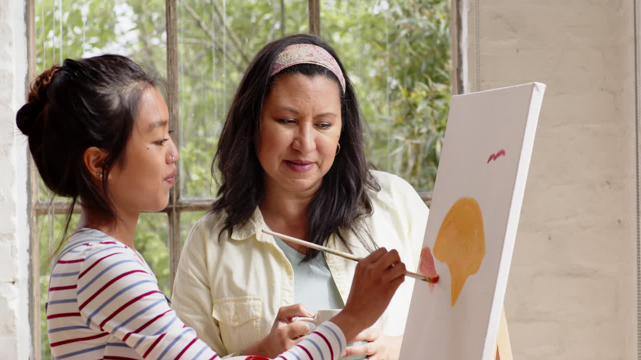 Multiracial young woman painting with grandmother, both smiling and enjoying creative time at home
