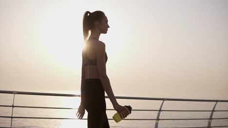 Gorgeous-side-view-of-a-pretty-girl-walking-on-the-road-along-the-sea,-the-sun-shines-quietly.-A-girl-in-black-leggings-and-top.-Pony-tail.-Carefree