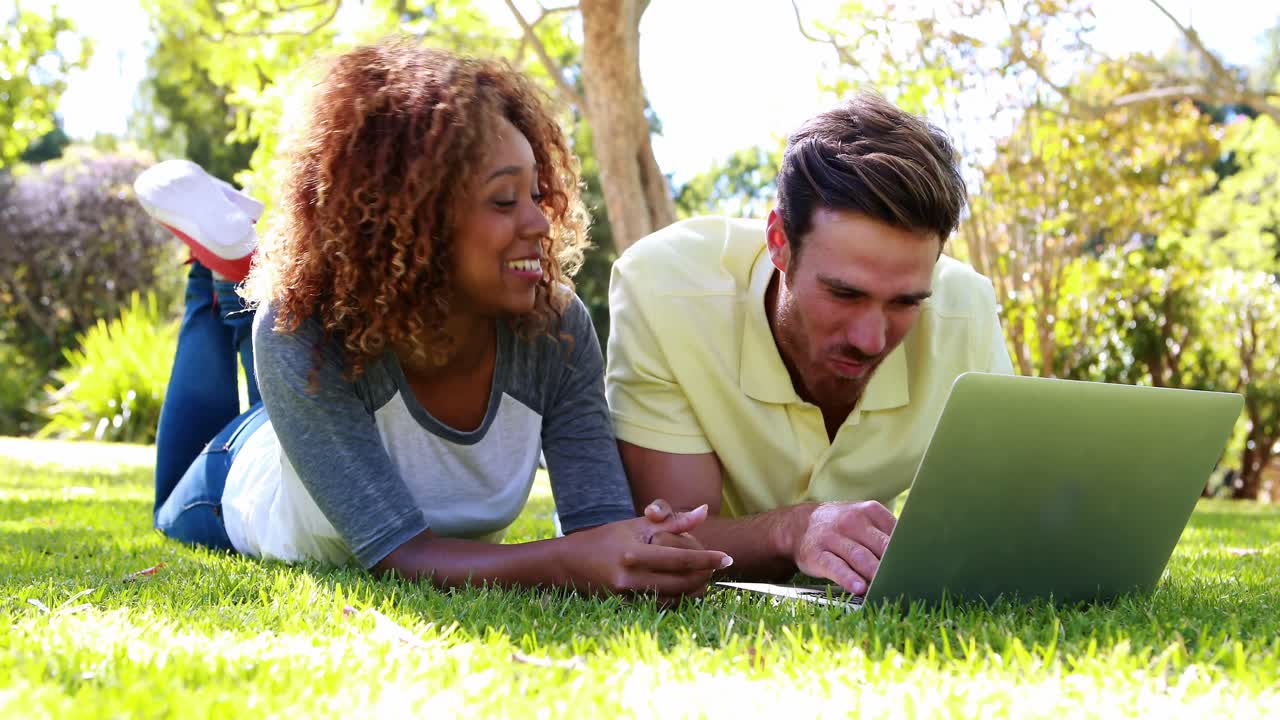 Couple lying on grass and using laptop in park