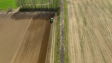 Aerial-shot-of-a-green-tractor-plowing-a-brown-field,-creating-distinct-patterns