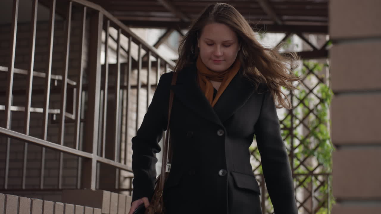 검은색 코트를 입은 젊은 여성이 차분한 표정으로 덮개가 있는 통로를 걸어 내려가고, 산들바람에 머리카락이 흩날립니다. 부드러운 햇살, 철제 격자, 푸른 잎이 뒤에 보이는 격자가 보이고 목적 있는 발걸음이 느껴집니다.