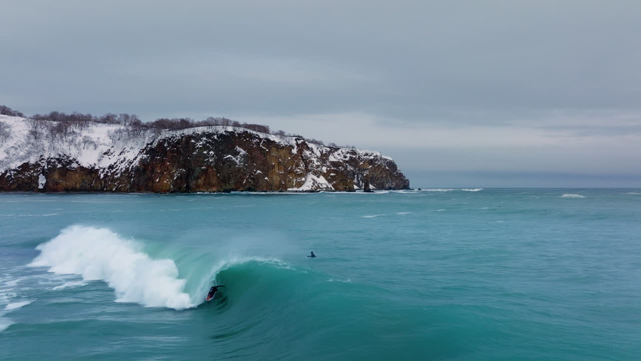 Surfař jedoucí na oceánské vlně blízko skalnatého zasněženého pobřeží, střední letecký sledovací záběr s oddálením, zatímco projíždí tunelem vlny.
