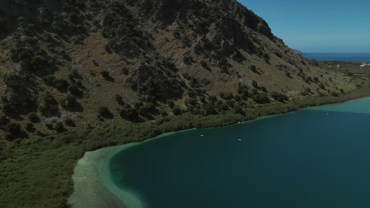 Rocky hills with green vegetation lead down to the still turquoise waters of Lake Kournas with natural shoreline formations and scattered trees visible
