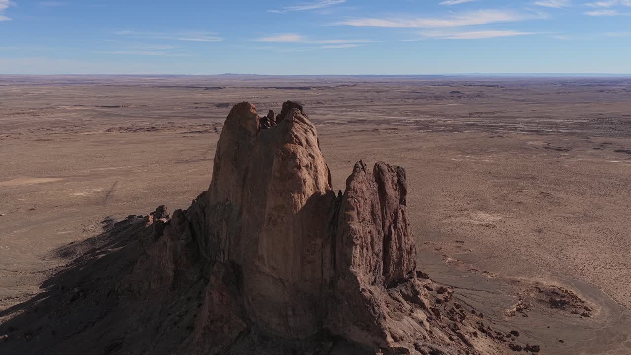 Una órbita aérea estable de dron captura el monolito aislado de butte de arenisca dentro de un vasto paisaje desértico árido, con formaciones geológicas escarpadas bajo un cielo azul brillante y pintoresco.