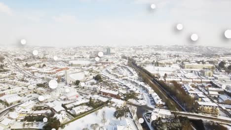 Composición-Digital-De-Múltiples-Puntos-Blancos-Flotando-Contra-La-Vista-Aérea-Del-Paisaje-Urbano-Cubierto-De-Nieve