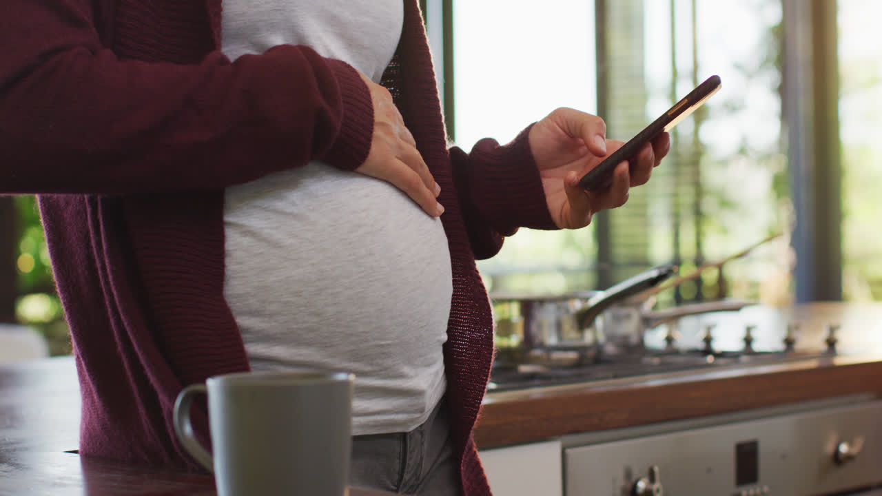 Midsection of caucasian pregnant woman touching belly and using smartphone
