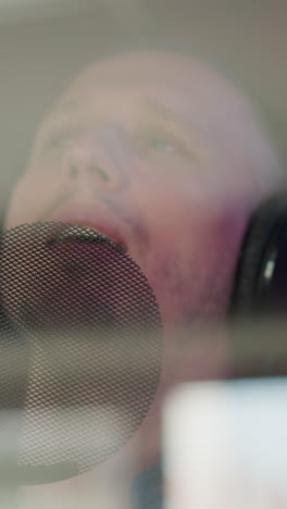 primer plano de un hombre cantando en un micrófono mientras lleva auriculares de estudio negros. su rostro está parcialmente oscurecido por el filtro pop del micró fón, capturando la intensidad y el enfoque de su actuación