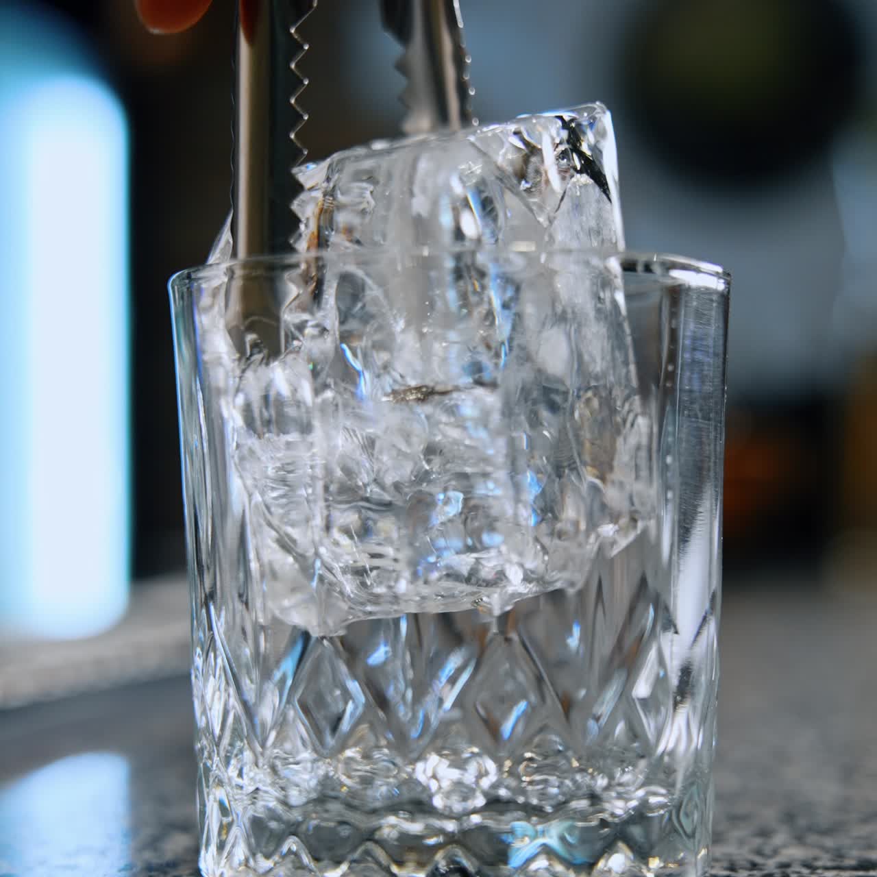 Een groot ijsblokje wordt met een pincet in een glas geplaatst. Het drankje wordt bereid in de bar. Close-up. Verticaal scherm. Wazige achtergrond