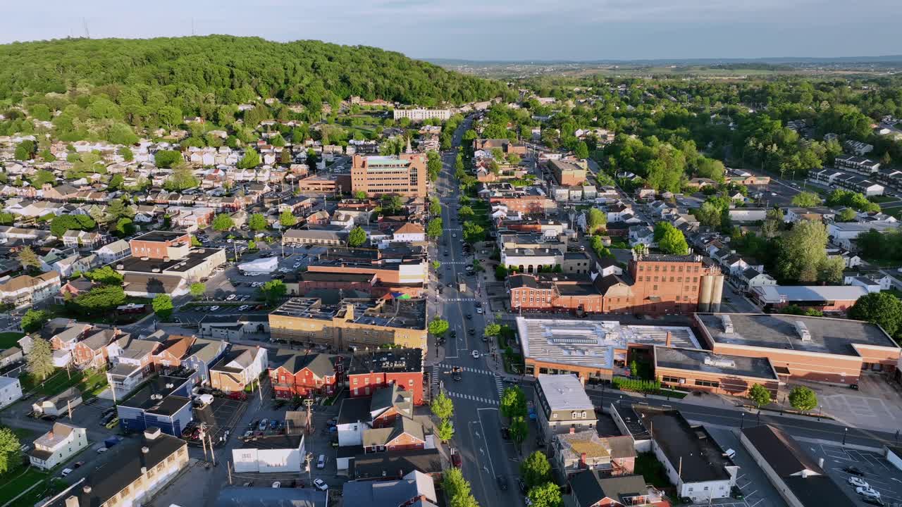 Main street junction of american town during sunny day in Ephrata, Pennsylvania. Green blossoming trees in hill in Background. Cars on street in downtown. with red brick buildings.
