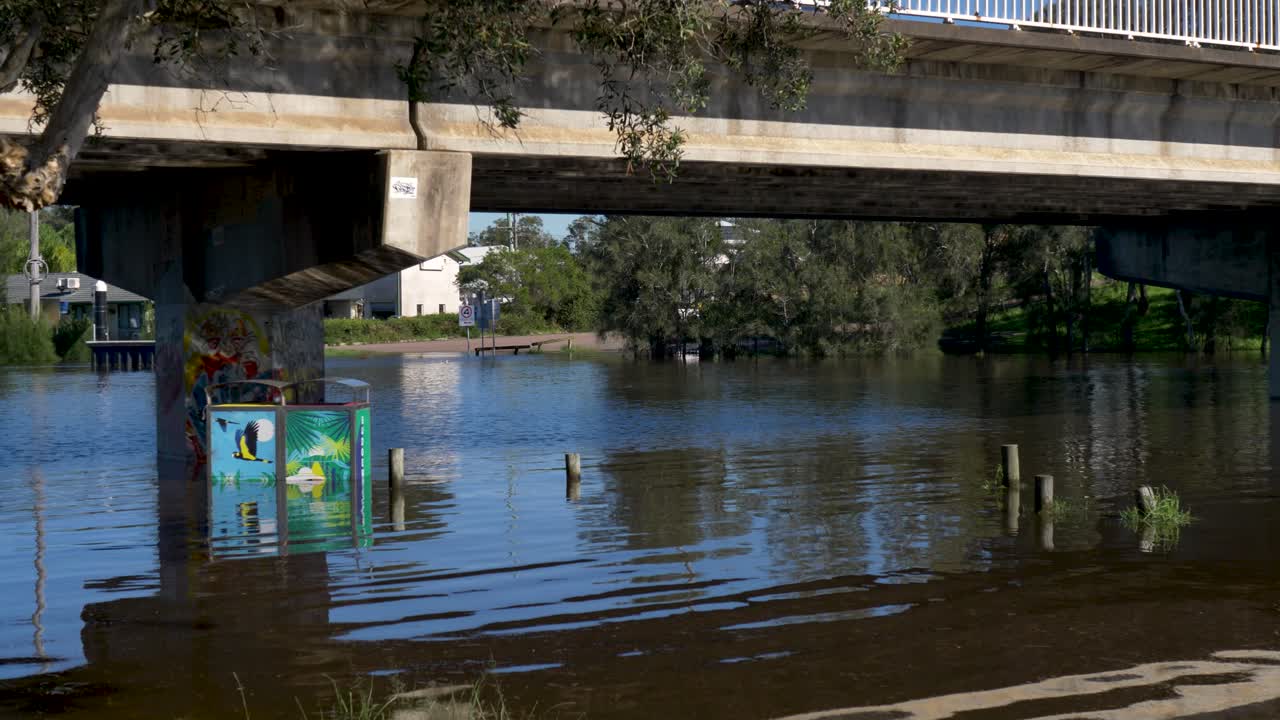 Slow motion landscape of two rubbish trash bins in flood water under Budgewoi creek and bridge lake underpass in park Central Coast Australian community hazard disaster risk weather event community