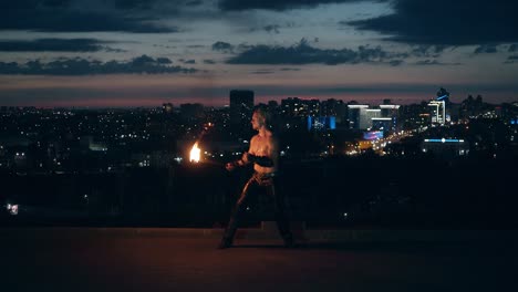 young blond male does tricks with fire breaths fireball in the middle of the night with city skyline in background 1