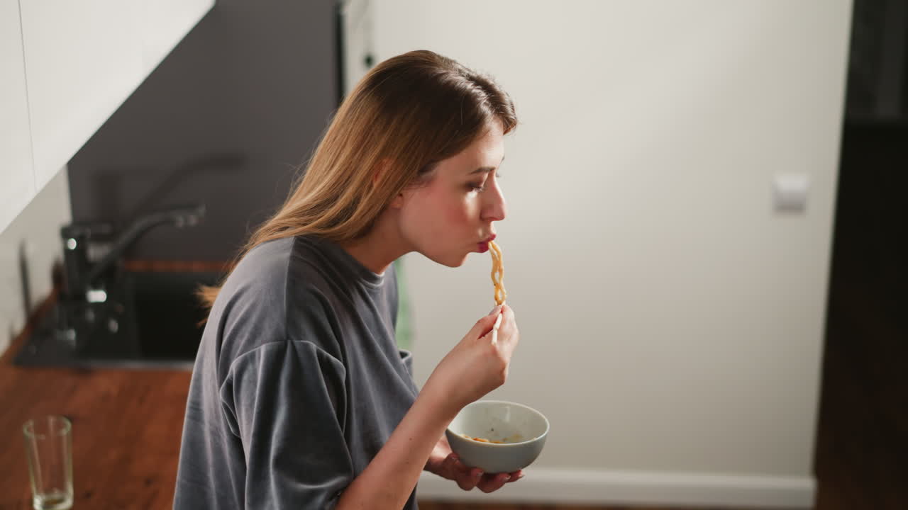 Side view of housewife eating breakfast standing in bright kitchen, soft light reflecting on face, holding bowl and chopsticks, empty glass cup on wooden counter behind her, casual morning scene