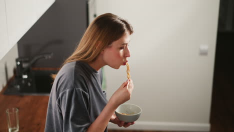 side view of housewife eating breakfast standing in bright kitchen, soft light reflecting on face, holding bowl and chopsticks, empty glass cup on wooden counter behind her, casual morning scene