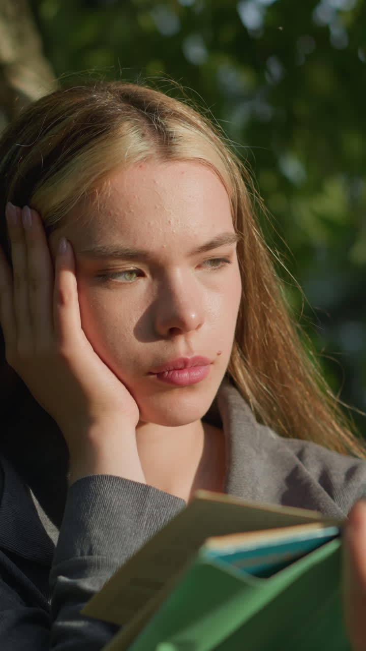dama de ropa gris sentada bajo un árbol con la cabeza apoyada en su mano derecha, sosteniendo un libro abierto mientras la luz del sol ilumina suavemente su rostro, parece profundamente pensativa