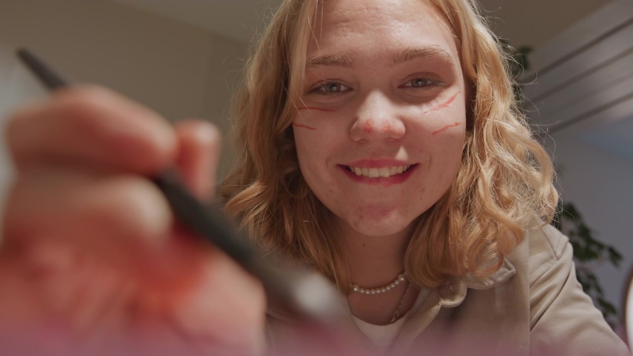 Close up of young woman applying makeup with steady hand indoors. Woman holds and uses various cosmetic tools focusing on precise application techniques against soft lighting. Background