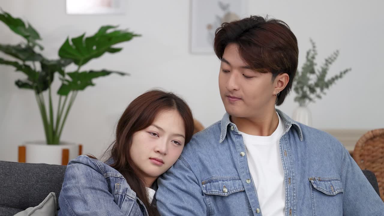 A young Asian woman sleeps on a man's shoulder while sitting on a sofa