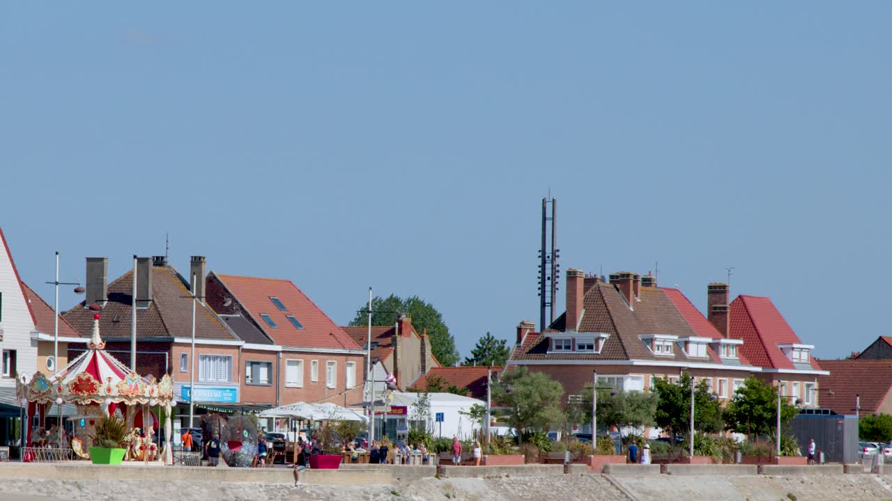 Colorful carousel rotates near beach, residential buildings in background, bright daylight, static wide shot