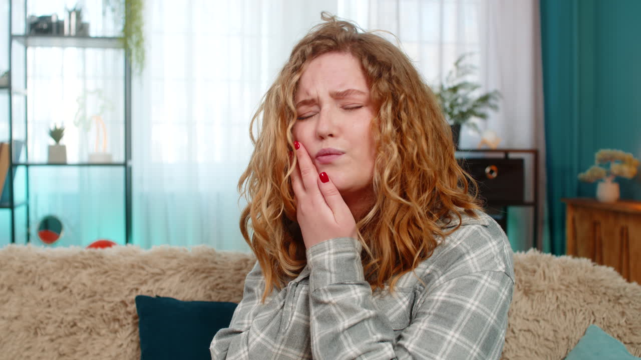 Woman sitting on sofa holding cheek with hand suffering from sharp toothache feeling pain and stress