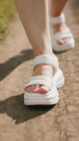 색 샌달을 입은 여자가 길을 고, 바닥에 그림자를 던지고, 은 빛 아래 초록색으로 둘러싸인 배경에 다른 사람의 부분적인 시야를 가지고 있습니다.