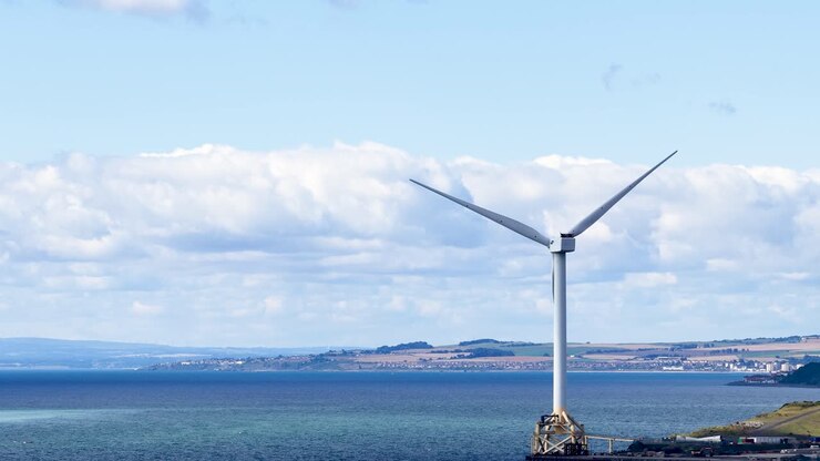 Large wind turbine spins steadily by the sea under bright daylight, wide static camera shot