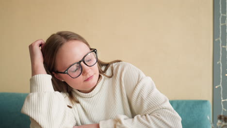 tired thoughtful woman in glasses wearing white sweater rests head on hand while seated near wall with visible socket and decorative string lights, expressing fatigue