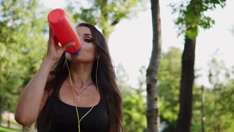 Sweating-woman-listening-to-the-music-and-drinking-pure-water-from-the-bottle-refreshing-herself-after-running-in-summer-park