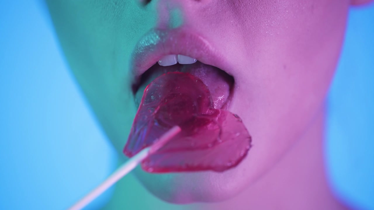 Primo piano della bocca di una donna che lecca un lecca-lecca rosso a forma di cuore, scatto in studio colorato