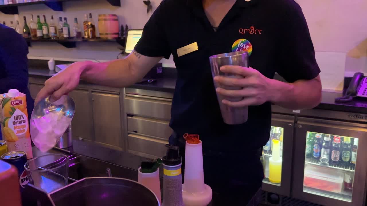 A bartender in a dimly lit Bangkok bar swiftly adds ice to a cocktail shaker, preparing a drink with efficient, practiced movements. Static camera, warm ambient lighting