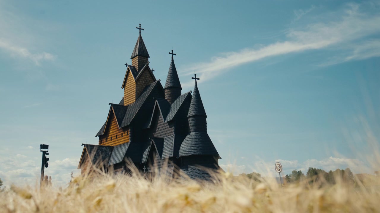 Heddal Stavkirke rejser sig over højt gyldent græs under en lys sommerhimmel i Telemark, Norge. Kompositionen fra lav vinkel fremhæver den karakteristiske trækonstruktion og den ikoniske middelalderlige silhuet