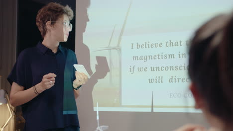 young female team leader standing by projection screen, giving presentation about wind farm and renewable energy to colleagues during night office meeting. over the shoulder shot